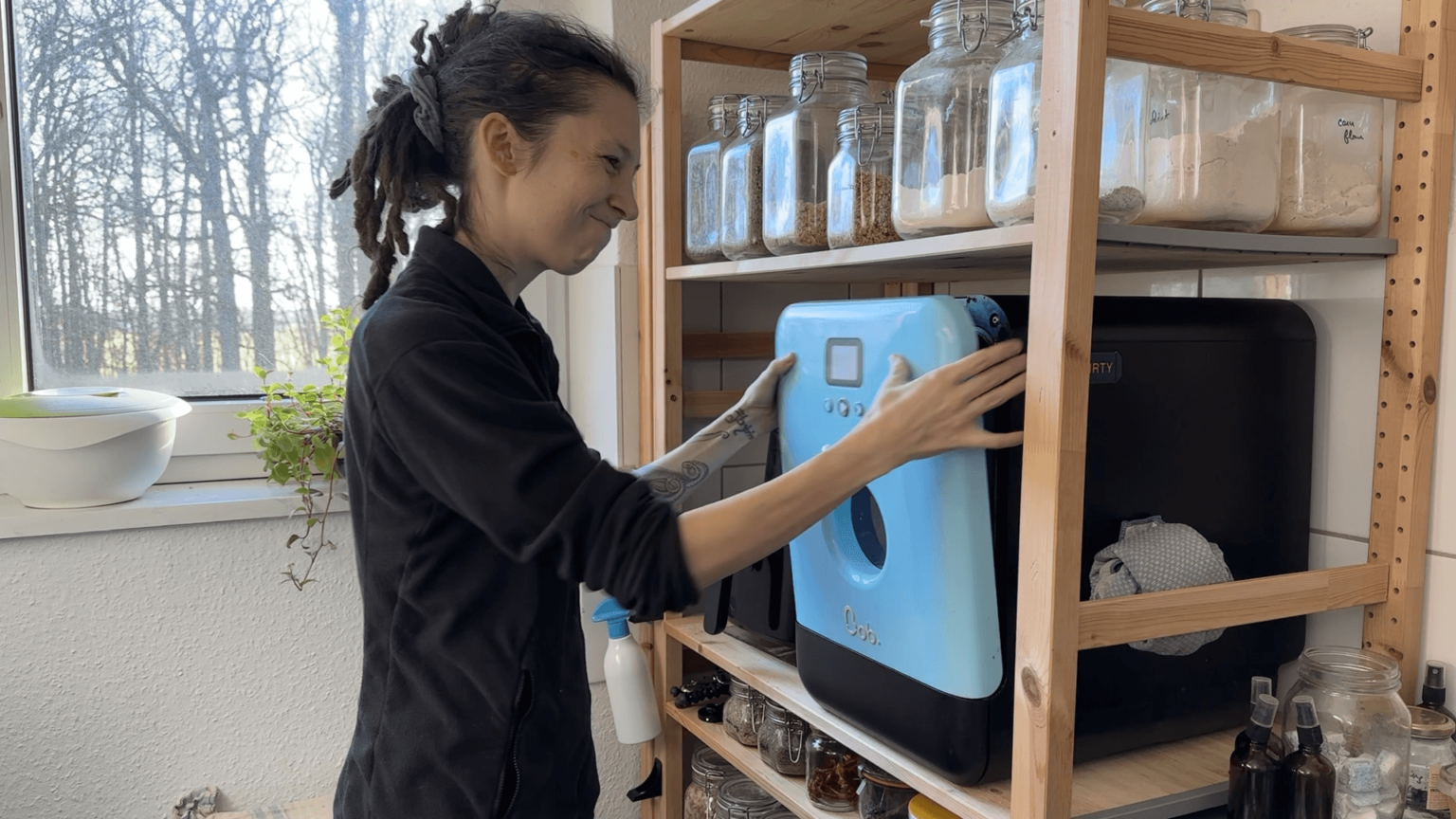 Kate looks very annoyed while trying to get the door of her dishwasher to stay closed. It's a turquoise table-top unit inside her kitchen shelf.