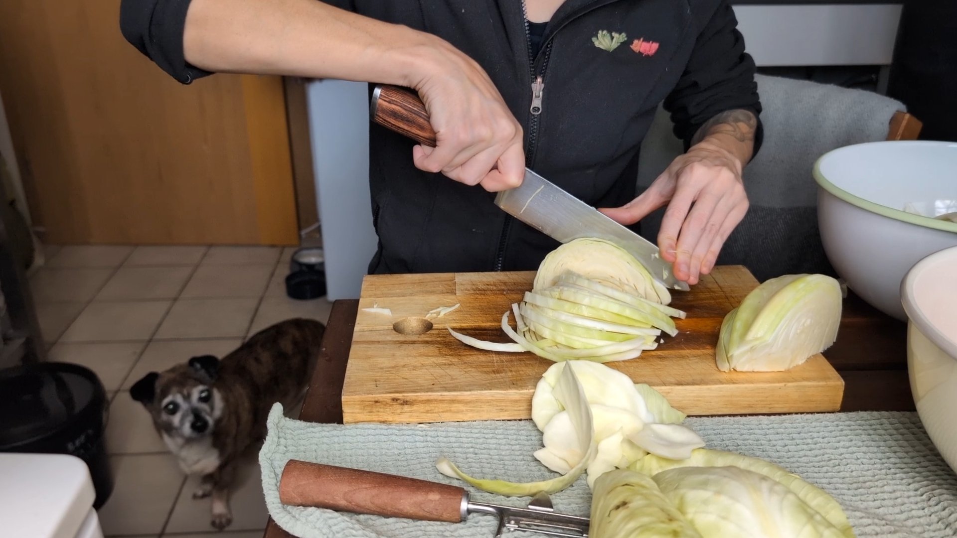 Kate sitzt an einem Holztisch und schneidet einen Kohl. Im Hintergrund ist ihr kleiner Hund Pepper zu sehen, der auf fallende Leckereien hofft.