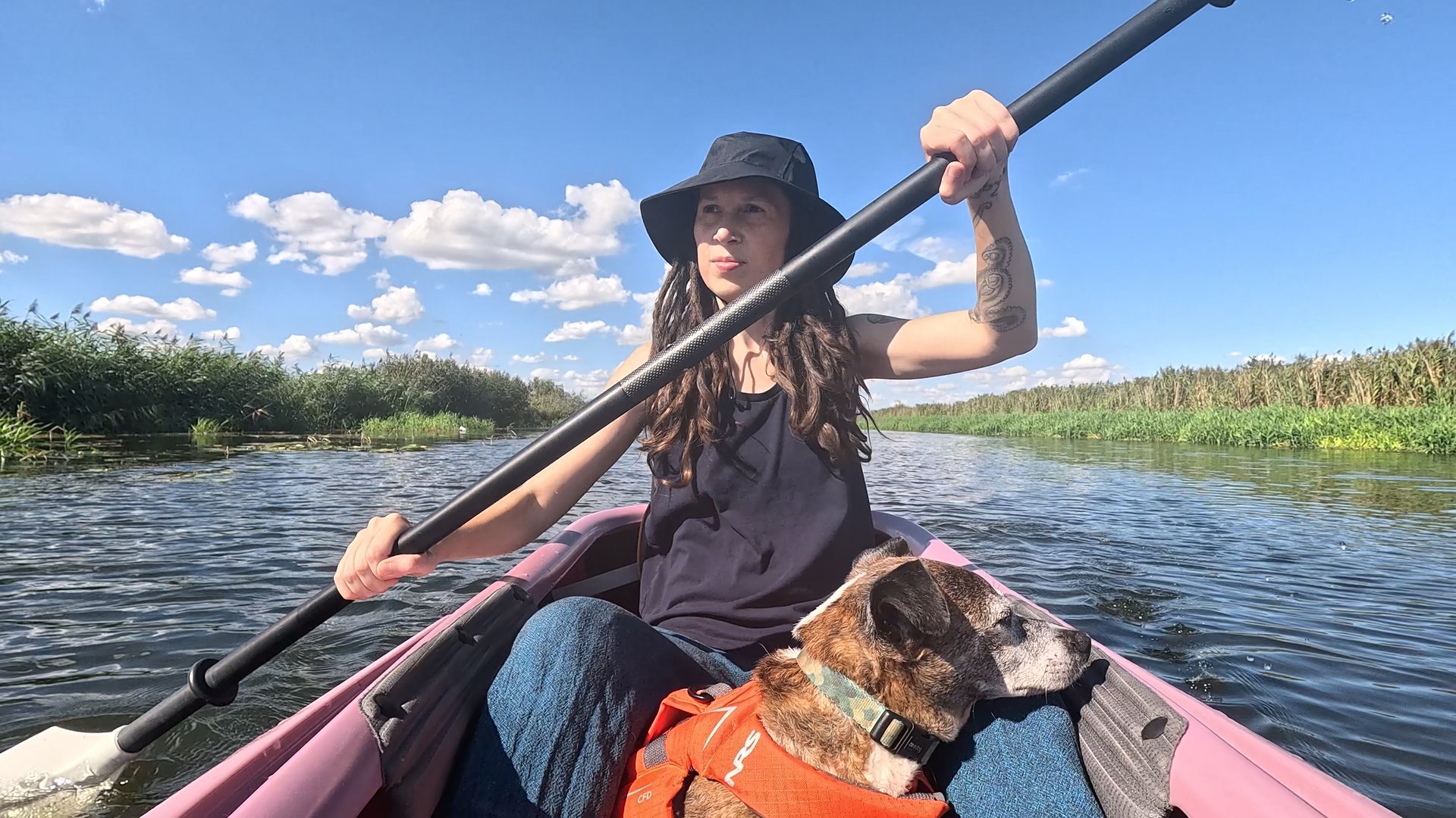 Kate in a kayak with Pepper on her lap. She is paddling on a river with reeds growing along the banks.
