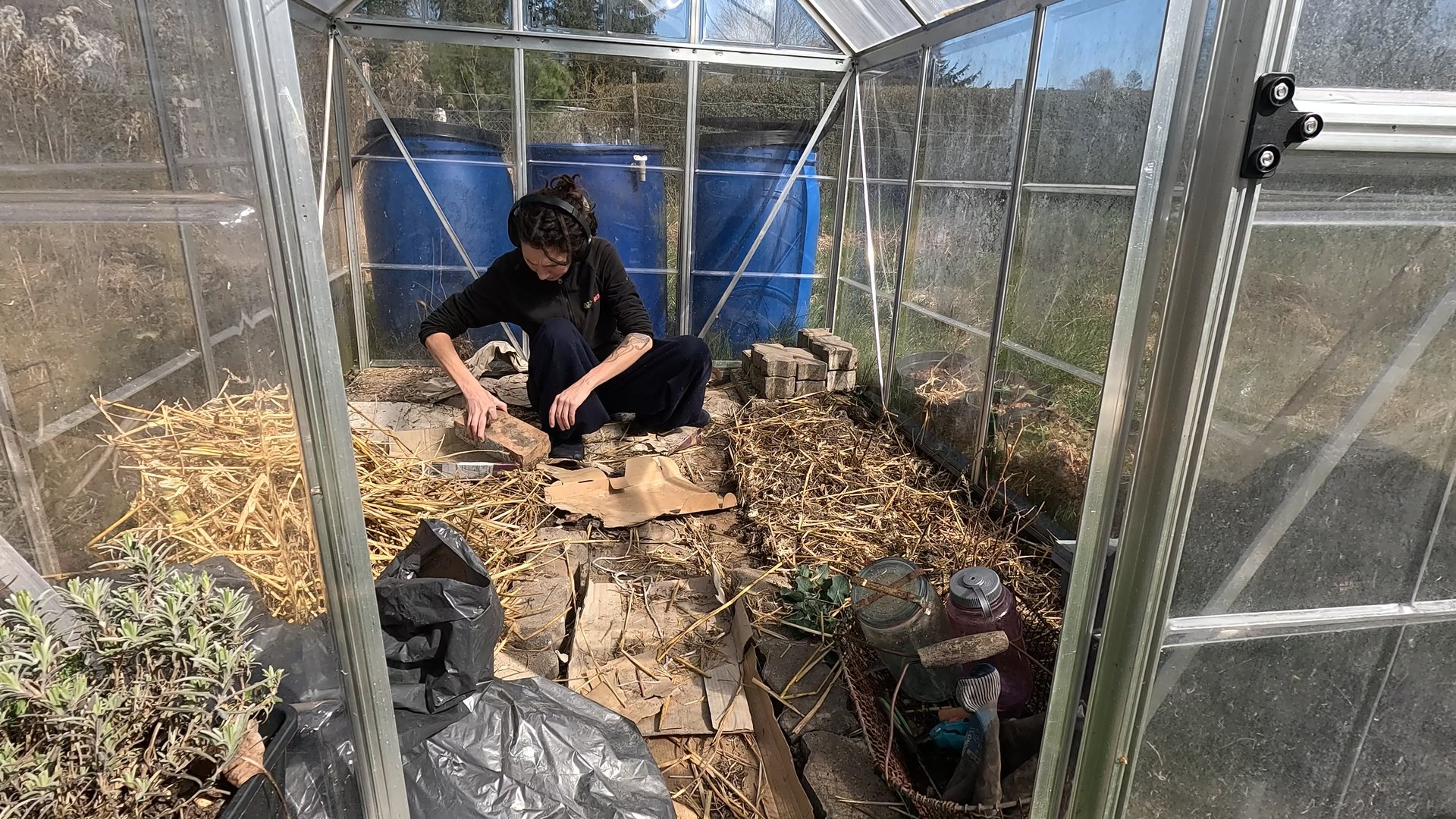 Kate is squatting in her greenhouse moving the bricks that used to be bed edges to stacks in the back corner.