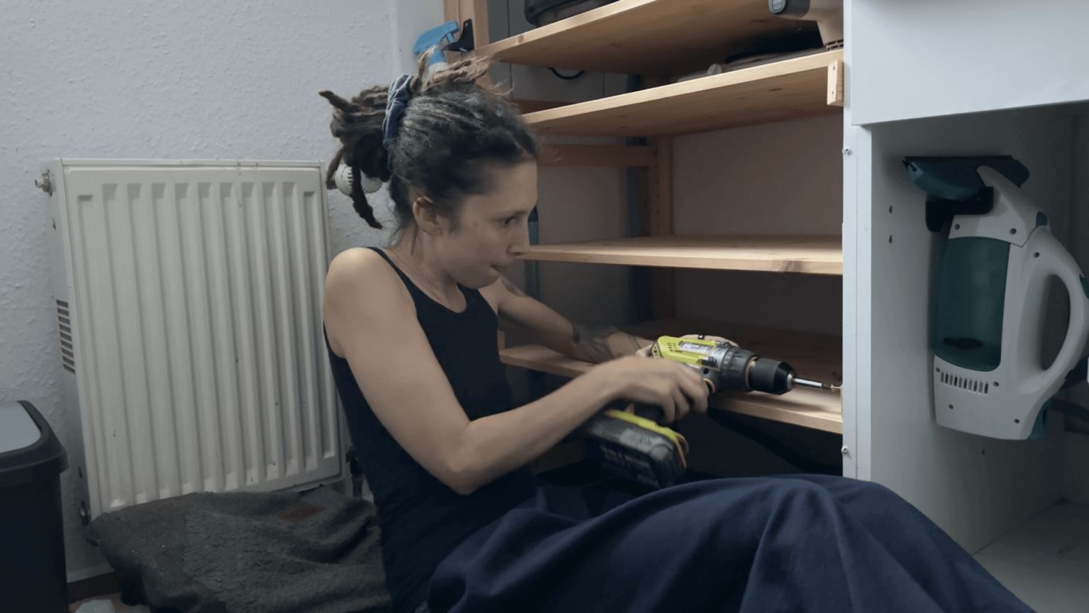 Kate is sitting on the floor, leaning back to screw in a board. She has her tongue between her lips in concentration.