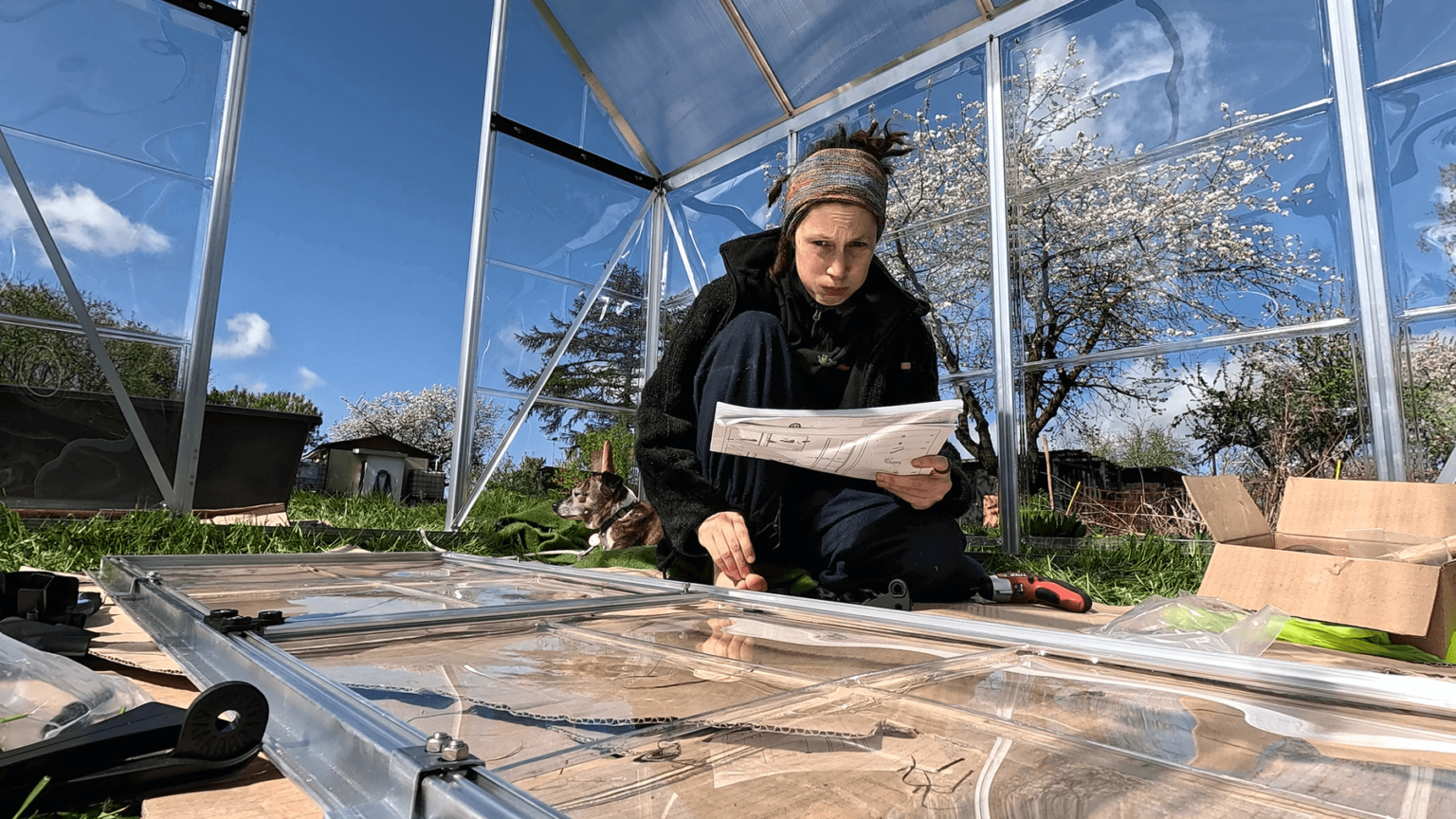 Kate sits in the middle of her new greenhouse, trying to understand the assembly instructions or the door. The door is on the ground in front of her. She is blowing air out in frustration.