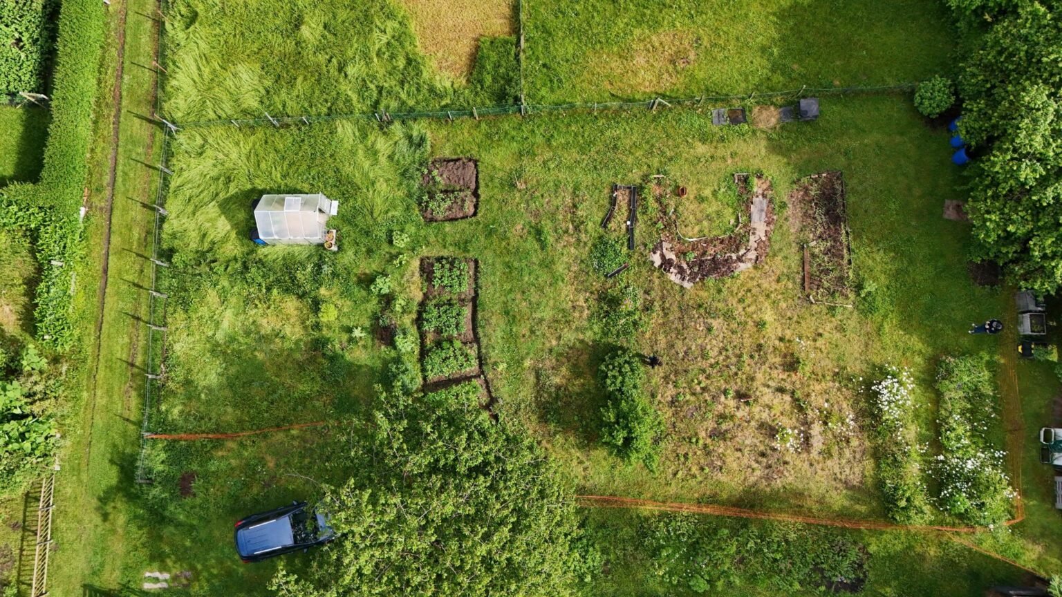 A drone image from above showing Kate's growing plot. On the left is a path, then the garden house, followed by the potato beds in a row from top to bottom. Further to the right, her tomato bed is forming a U-shape next to the traditionally shaped onion-and-garlic bed. Two flower strips are on the bottom right.