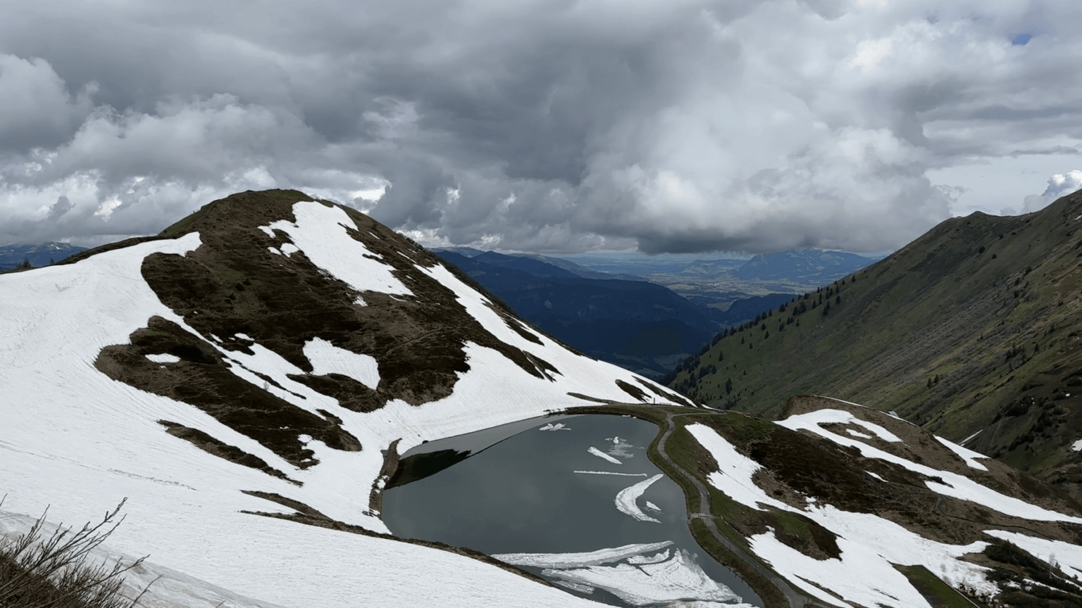 Drone footage showing a peak partially covered in snow and an artificial lake. A path runs past the lake. In the background, a valley is visible but clouds cover most of the mountainside.