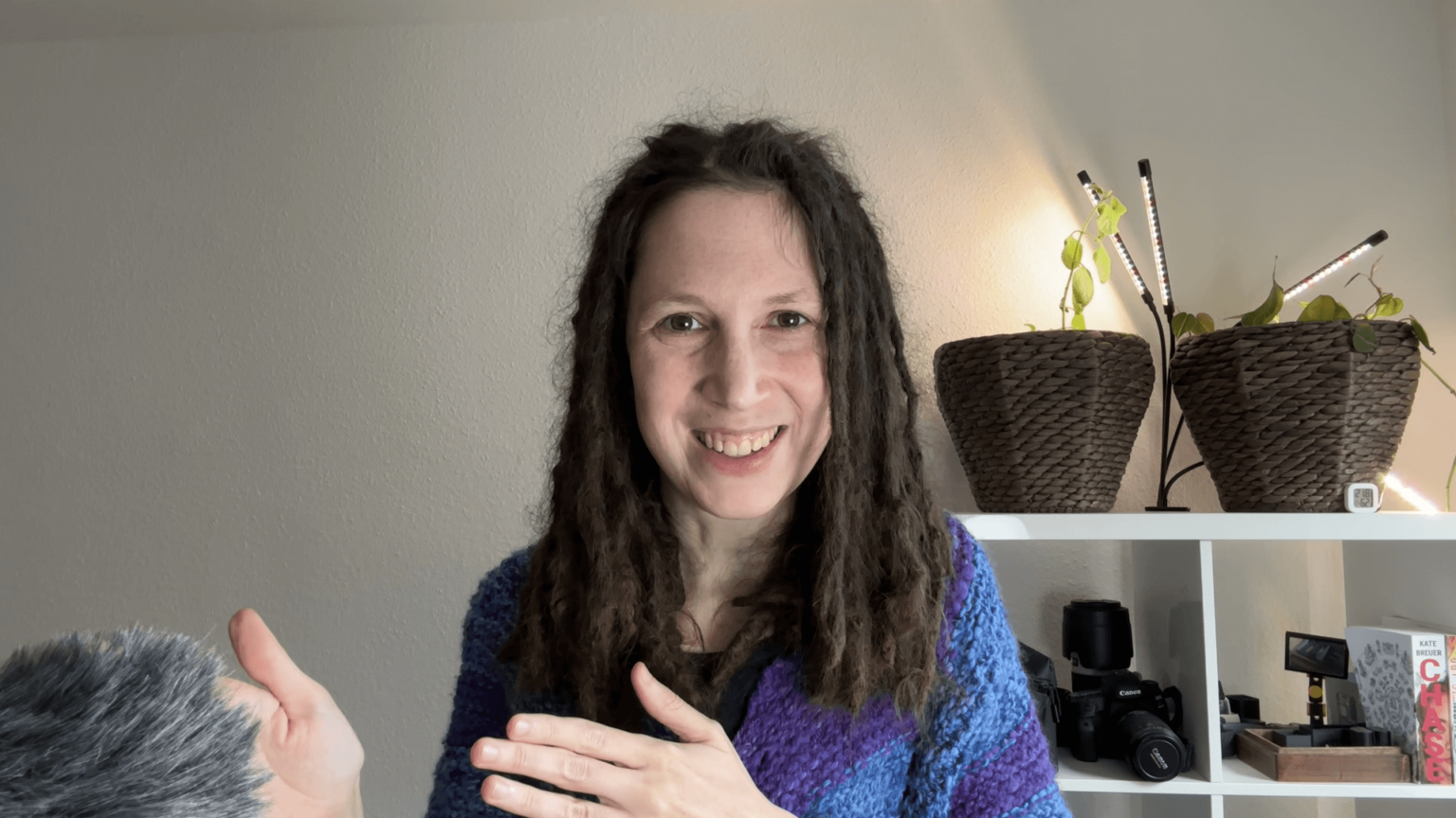 Kate looks straight at the camera. A microphone is just visible at the edge of the screen. Behind her, are some plants and camera equipment on a shelf. She is smiling. Her hands are held up in a way that indicates she is likely speaking.