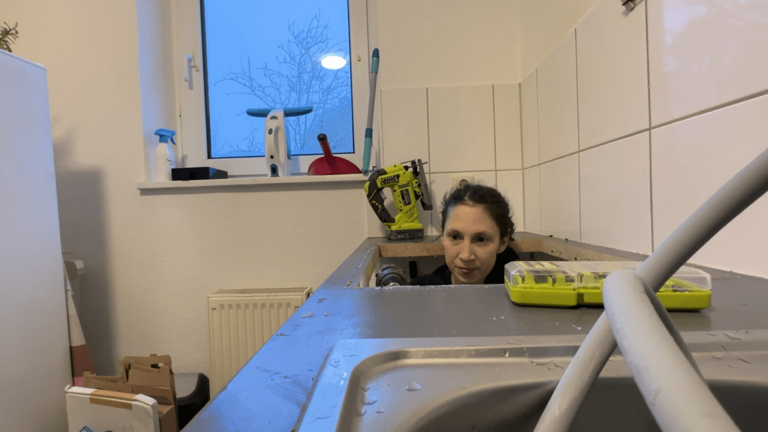 Kate looks straight at the camera. A microphone is just visible at the edge of the screen. Behind her, are some plants and camera equipment on a shelf. She is smiling. Her hands are held up in a way that indicates she is likely speaking.