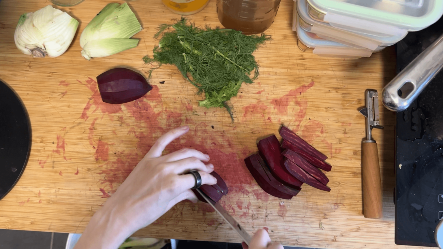 A view from above onto a wooden countertop. Kate cuts beetroot into slices. Thereare fennel greens and the rest of the fennel toward the top of the image and a stack of three glass containers.