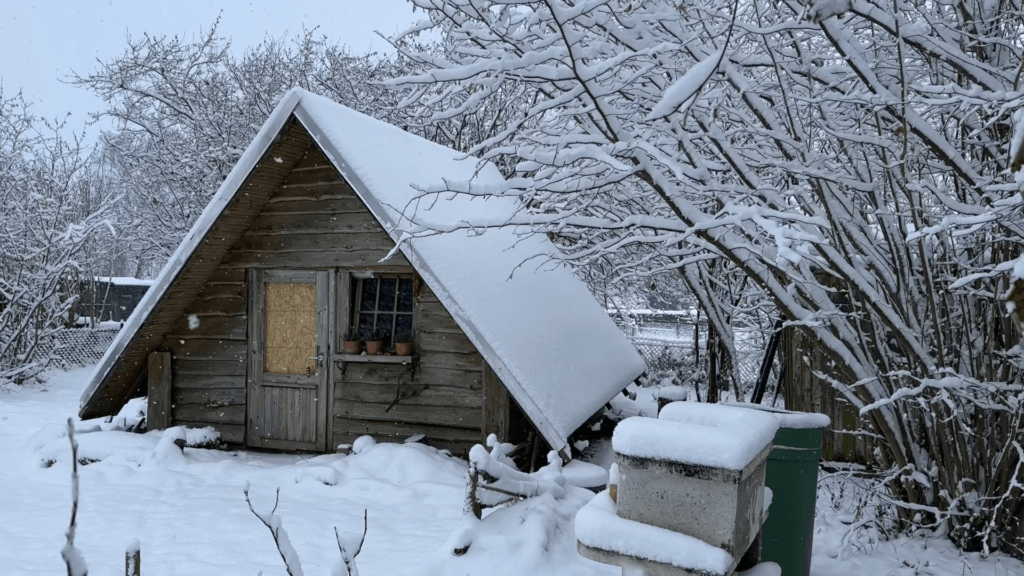 Kate's garden house is snowed in. Everything is covered in a blanket of white. Because the garden house is an A-frame the white roof almost reaches the white ground. Snow flakes are falling, only visible against the darker wood of the garden house front.