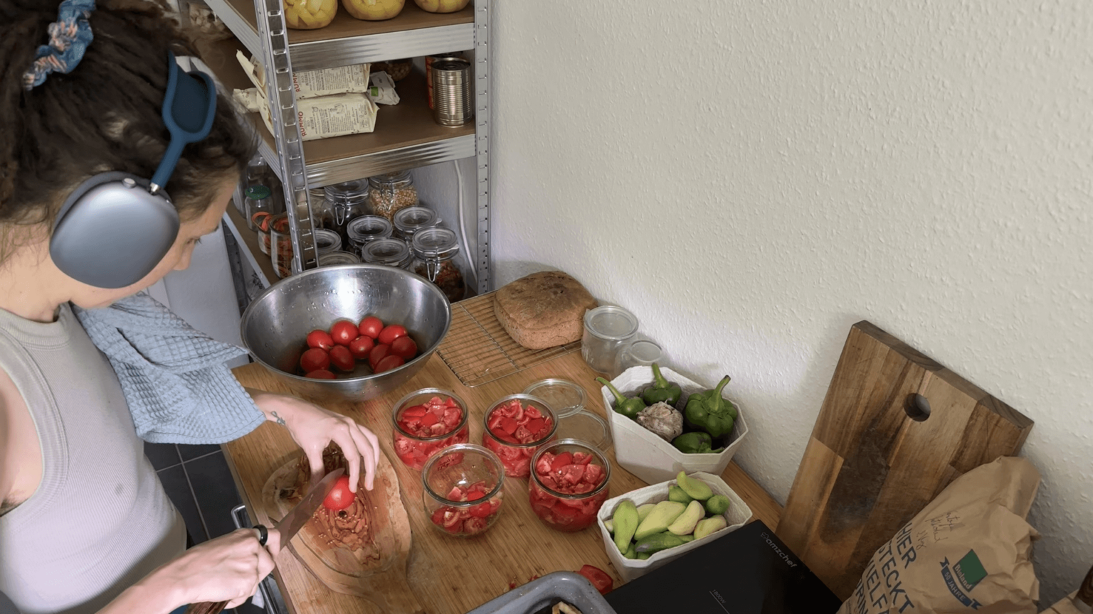 A view from above. Kate is wearing headphones yet again and cutting a tomato in half on a wooden board. On the counter are canning jars filled with tomato pieces, a bowl with washed tomatoes and some other vegetables.