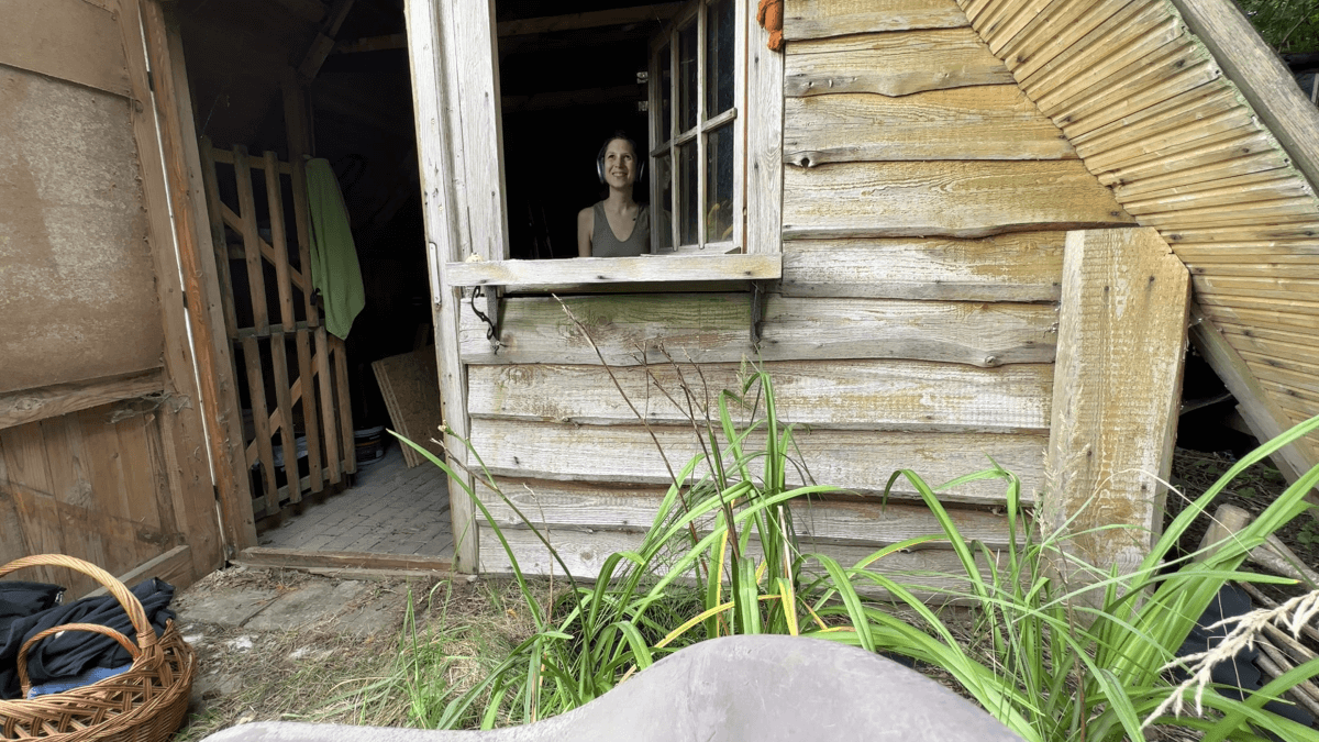 Kate stands inside the open window of her garden house. She is smiling.