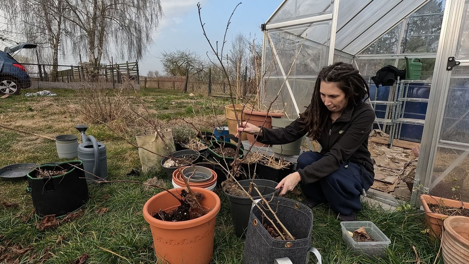 Kate is squatting in front of her greenhouse surrounded by pots holding branches. She is holding one branch to repot.
