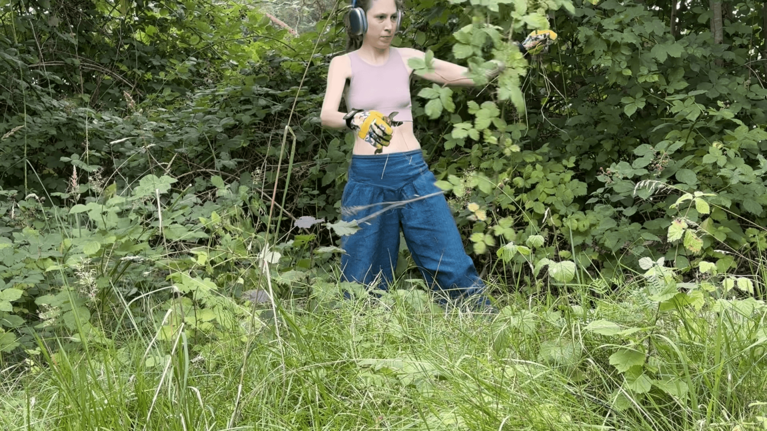 Kate holds apair of garden shears in one hand, a large blackberry bramble in the other. She is standing in the middle of a blackberry patch.