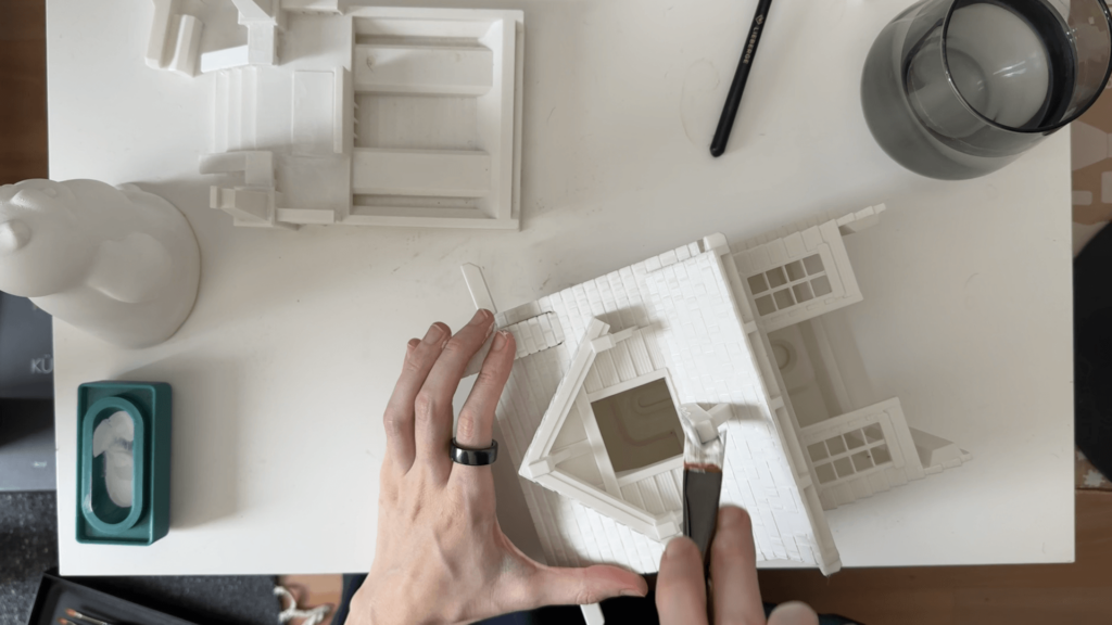 A view from above onto a white desk. Kate is painting a white bird house white.