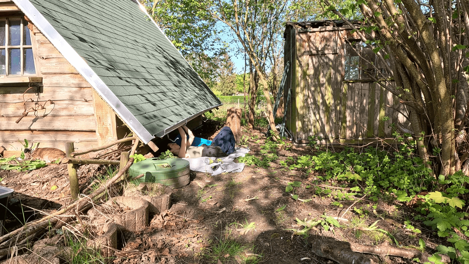 Kate lies on a blanket on the ground with her head under her A-frame hut's roof. She is installing the brackets for rain gutters. Toward the front of the garden house, a rain barrel is dug into the ground.