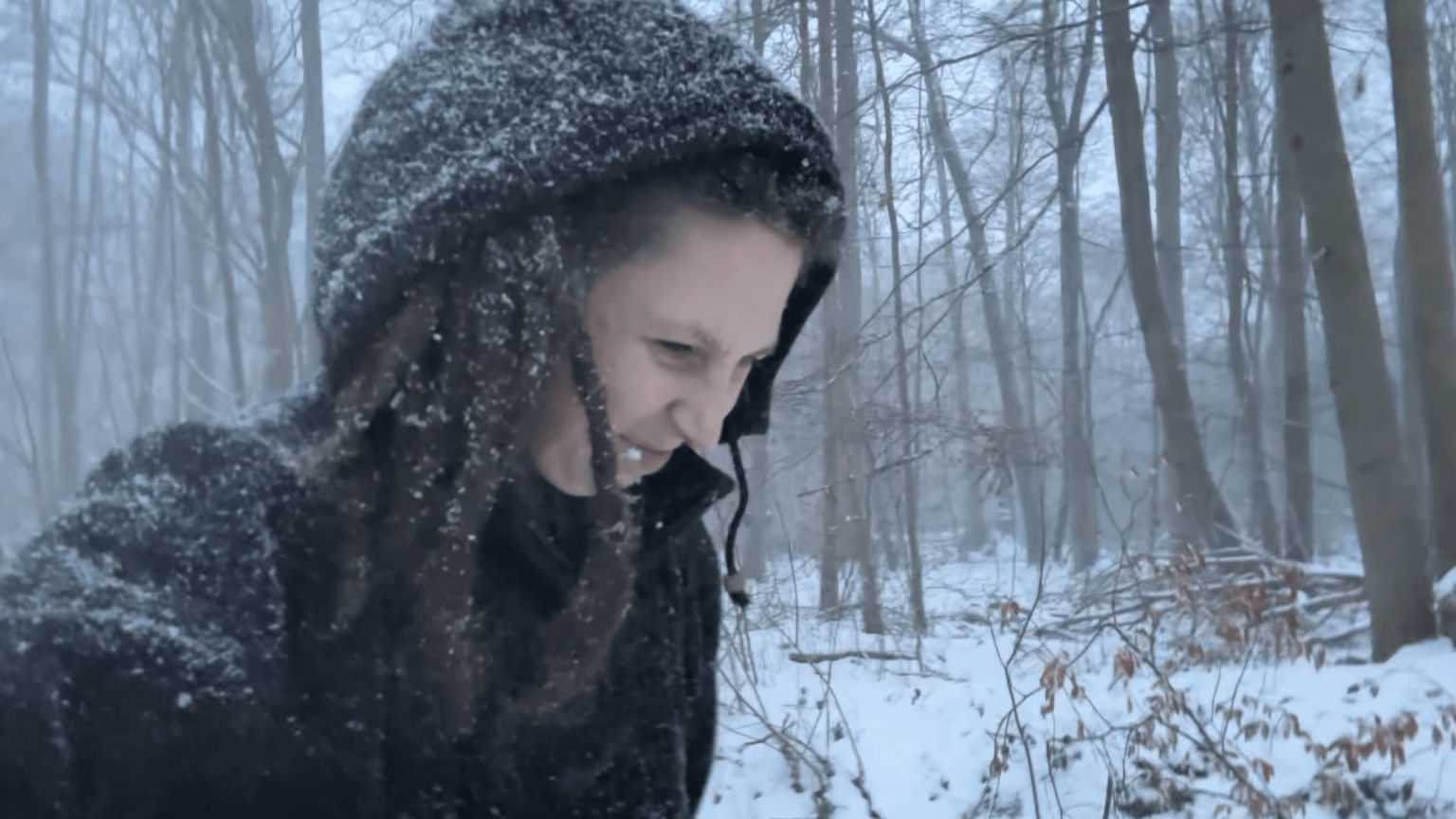 Kate is all covered up in athick winter jacket. Her jacket and dreadlocks are covered in snow. She is looking quite grumpy. Behind her, the forest is covered in snow.