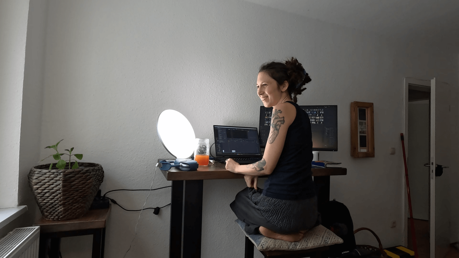 Kate kneels on her high bar chair in front of the bar table she uses as a desk. A daylight lamp is turned on on her desk. Two screens show the editing interface of DaVinci Resolve. She is looking to the left at the window, smiling.