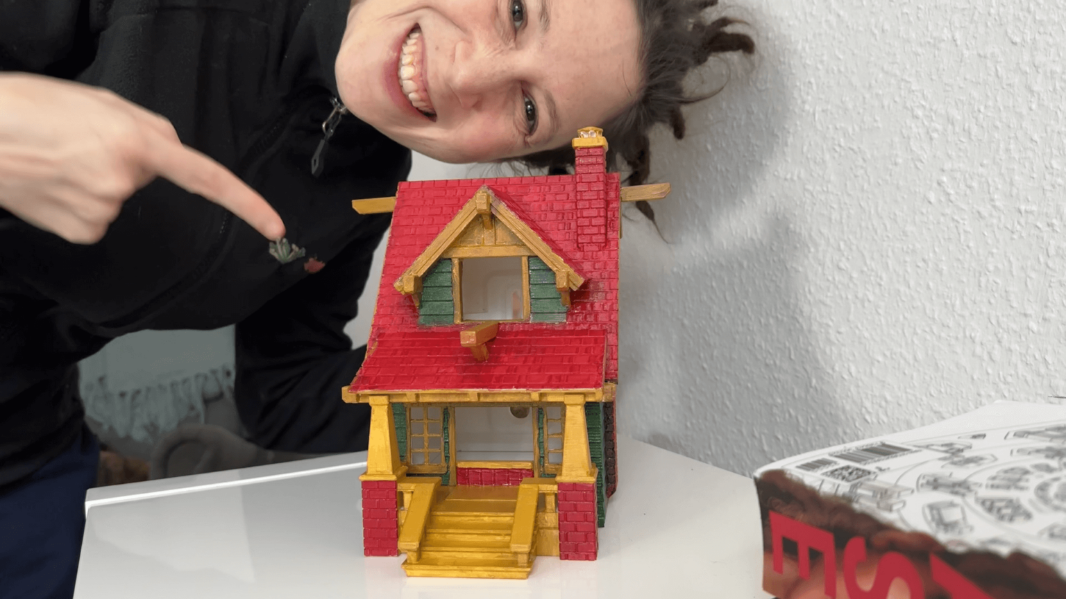 Kate points to a freshly painted colorful birdhouse. The birdhouse looks like a small house. She is leaning into the frame, grinning.
