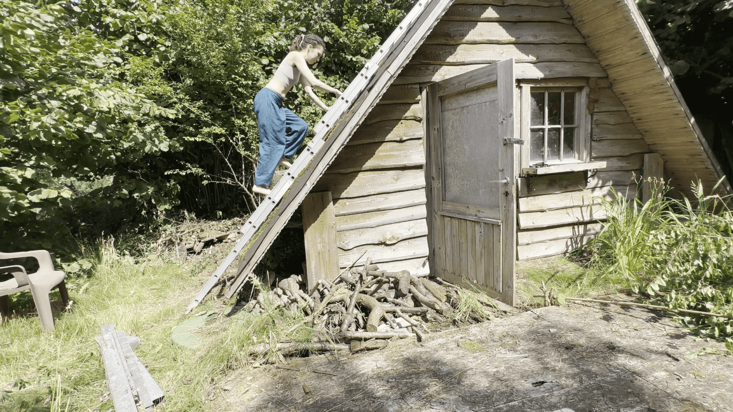 Kate climbs a ladder onto her garden house roof. The building is an A-frame building with the roof reaching to almost the ground.