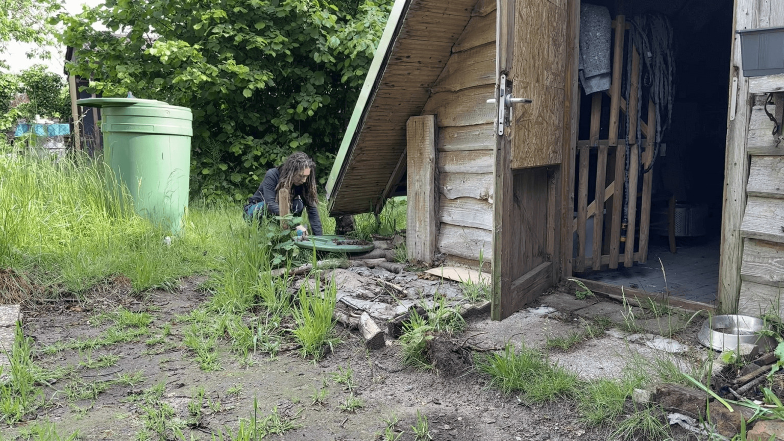 Kate squats next to a rain barrel dug into the ground next to her A-frame garden house. Another barrel stands behind her. The garden house door is open.