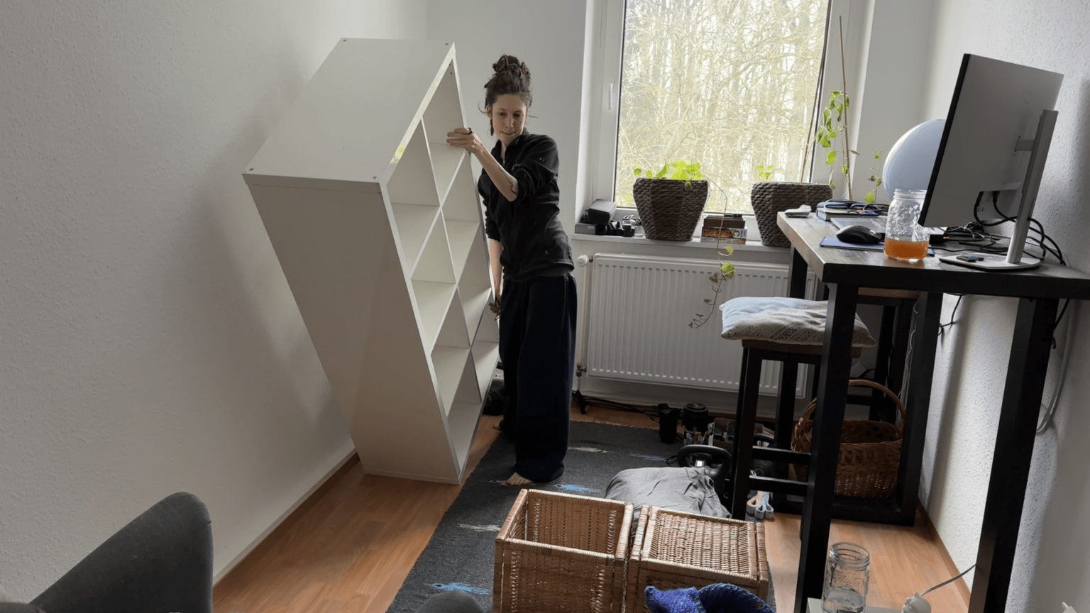 Kate is turning a shelf on its side. It's a white shelf with eight square boxes. There are items on the floor that belong into the shelf. She is on the left side of the room with a window behind her to her right and then a high desk with a bar chair.