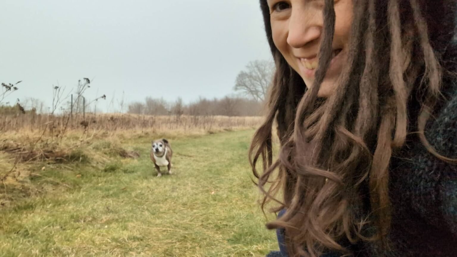 Kate lacht in die Kamera während ihr Hund Pepper im Hintergrund auf sie zurennt.