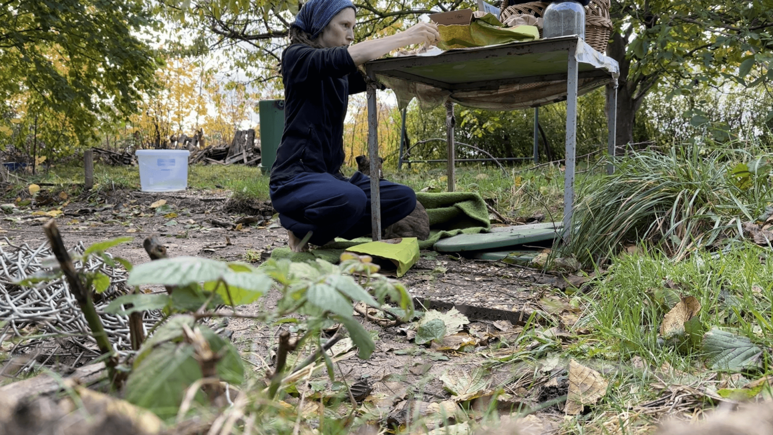 Kate squats at a metal table in her garden. She is peeling garlic. Her basket and some paper bags are on the table. Pepper is just visible next to her.