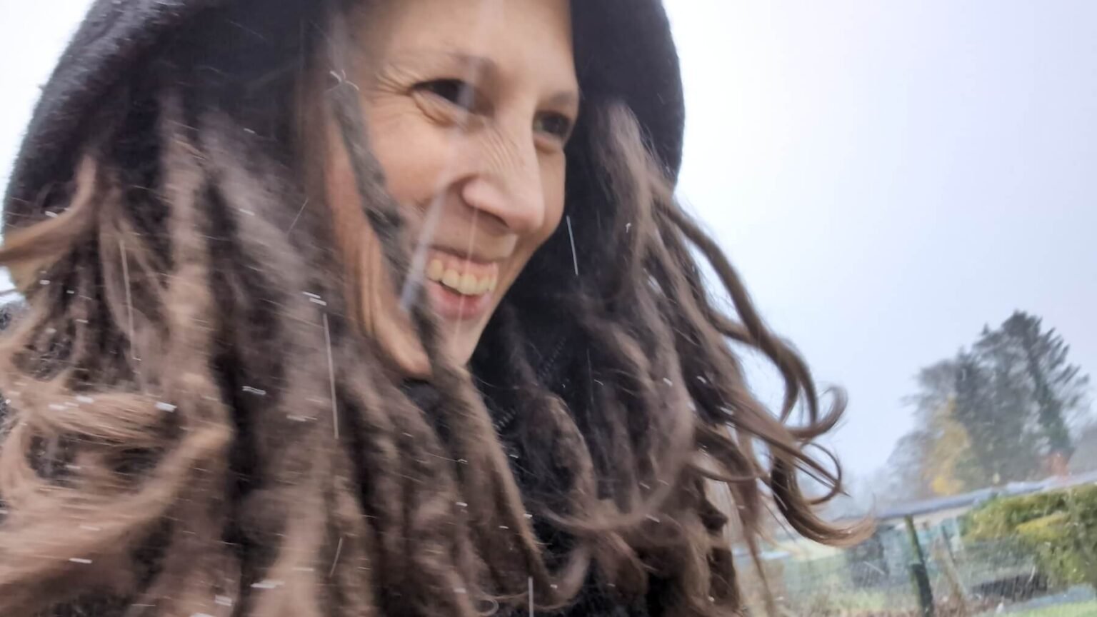 A portrait of Kate walking through dense snow. Snowflakes are visible as spots and stripes. Her dreadlocks stick out from her hood.