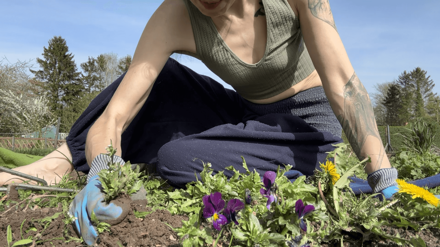 Kate sits on her legs next to a garden bed. She is pulling weeds by hand. Her face is out of frame. Her muscles show strain while pulling.