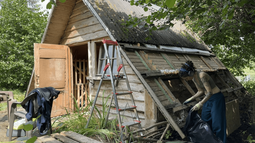 Kate brushes styrofoam that has disintegrated from a half-open roof. There is a ladder and quite a bit of greenery.