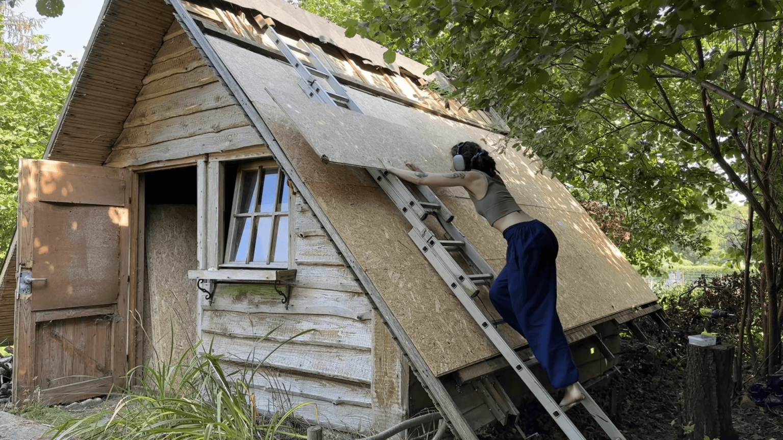 Kate pushes an OSB board up a ladder onto a roof that is already partially covered in wood.