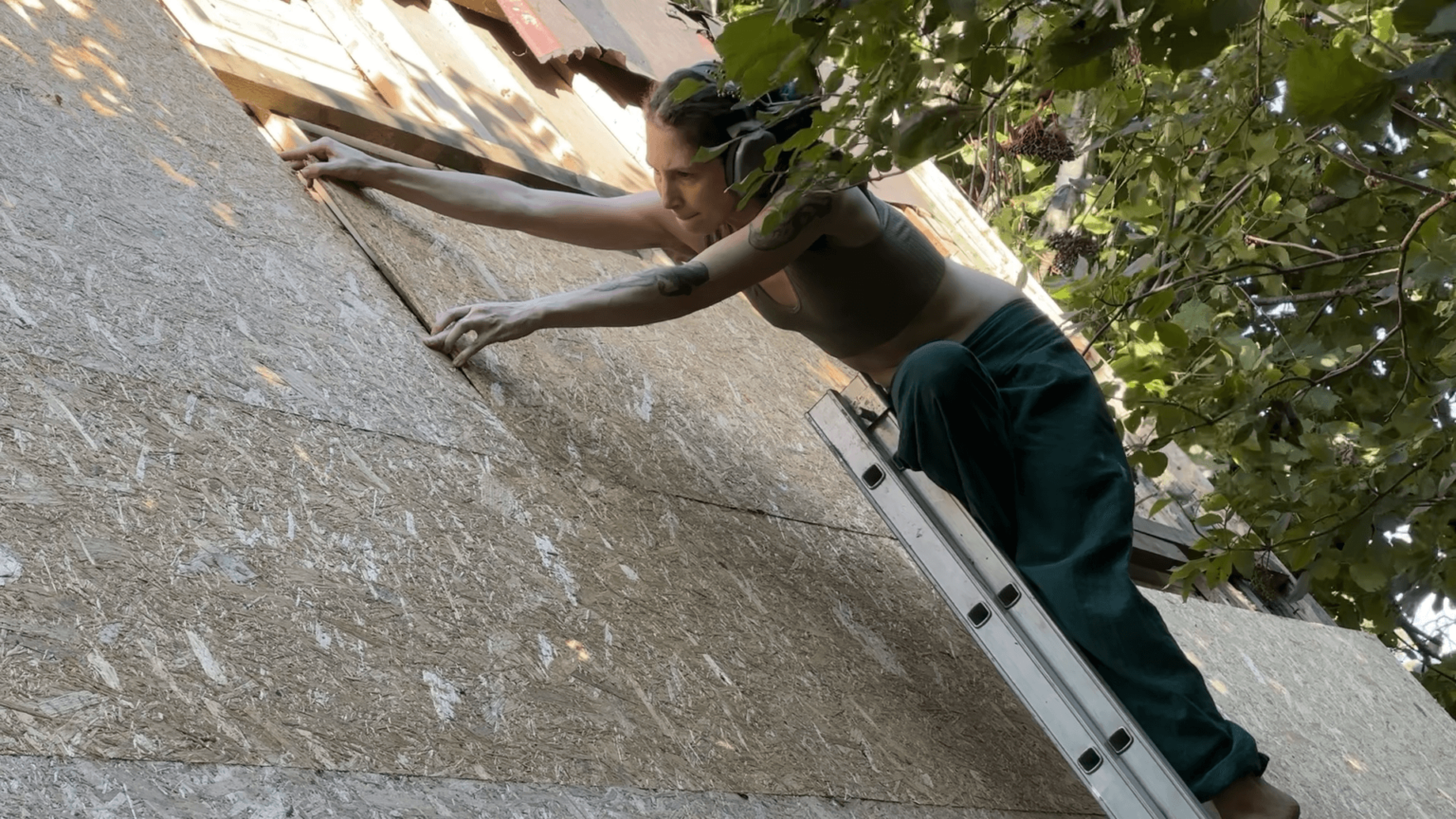 Kate stands on a ladder, leaning far to push a wooden board into the next one on the roof.