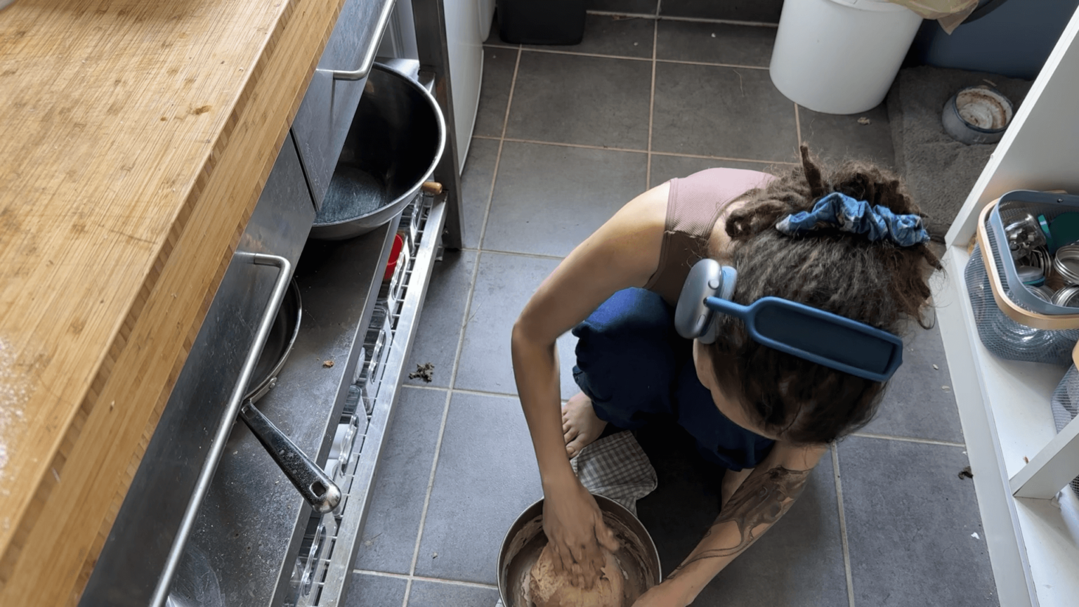 Kate kneels on the kitchen floor, working a sourdough bread in a metal bowl. The kitchen floor is dirty.