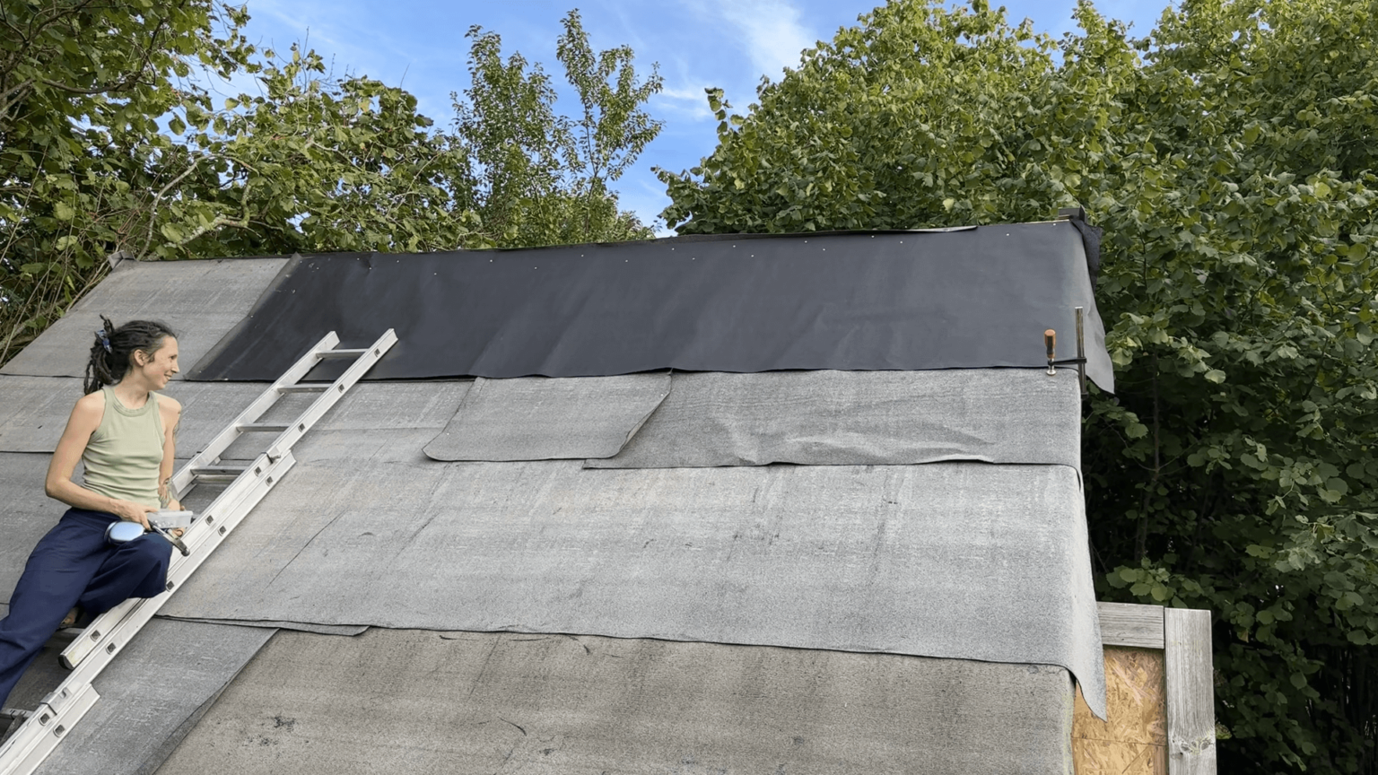 Kate sits on a ladder leaning against the roof of her garden house. The bitumen layer is on. It is two different colors.