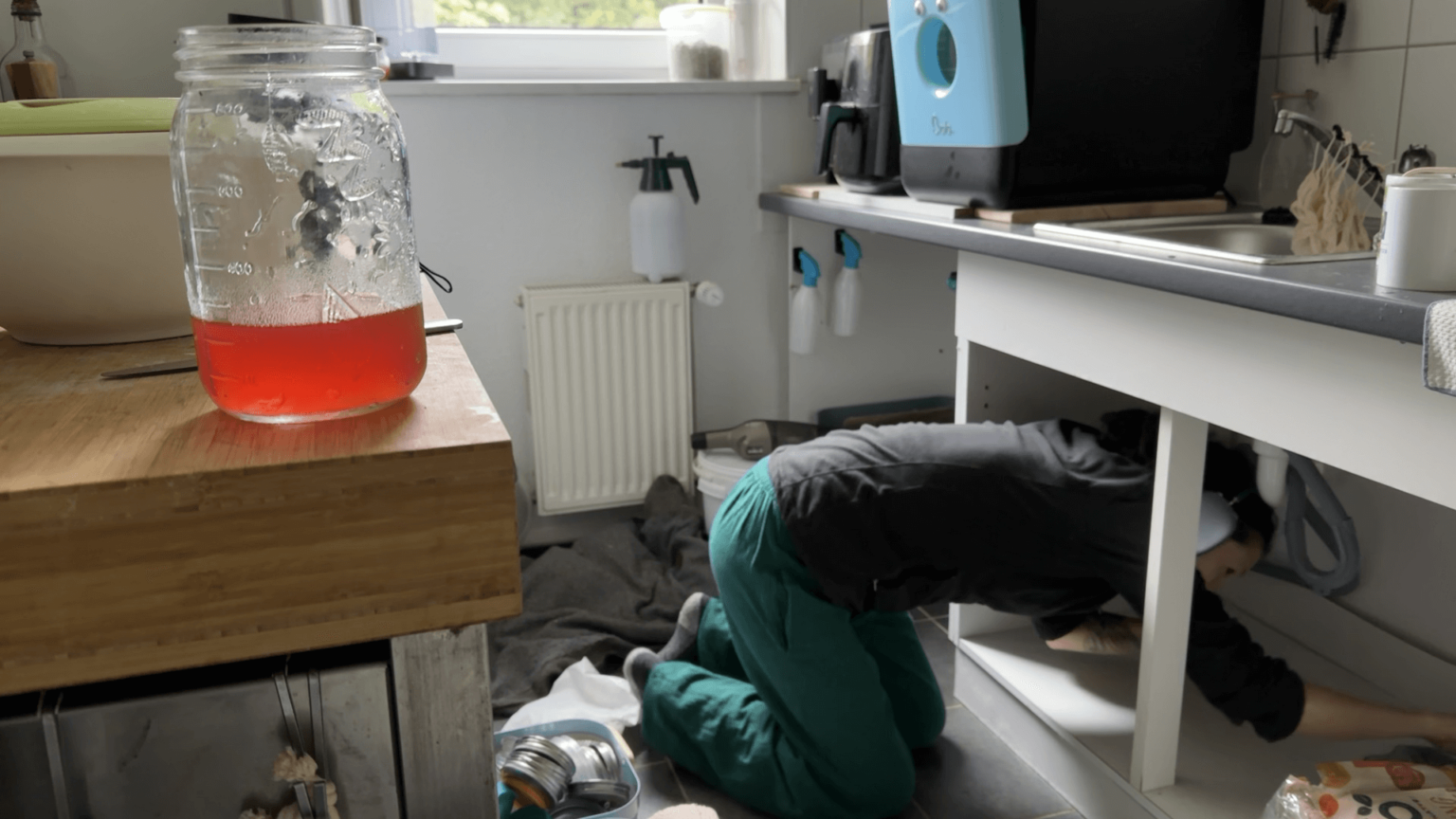 Kate is halfway into the cabinet under her sink. The cabinet does not have doors. It isn't fully clear from the picture but she is cleaning the cabinet with a rag. She is out of focus because the camera focused on a mason jar with reddish liquid inside.