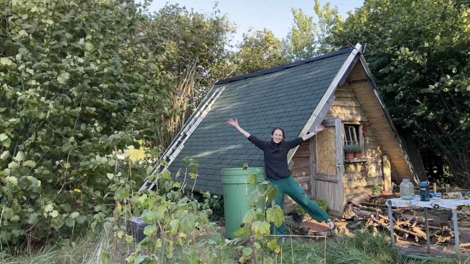 Kate with her arms and legs wide jumping in front of her finished garden house roof. The finishing layer of turquoise bitumen tiles is on.