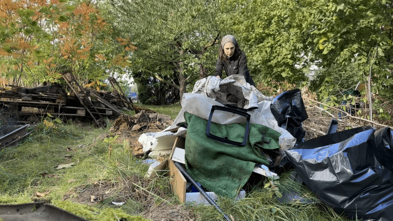 Kate stuffs a moldy tarp onto a large pile of random trash in her garden. There is another pile of broken pallets behind her.