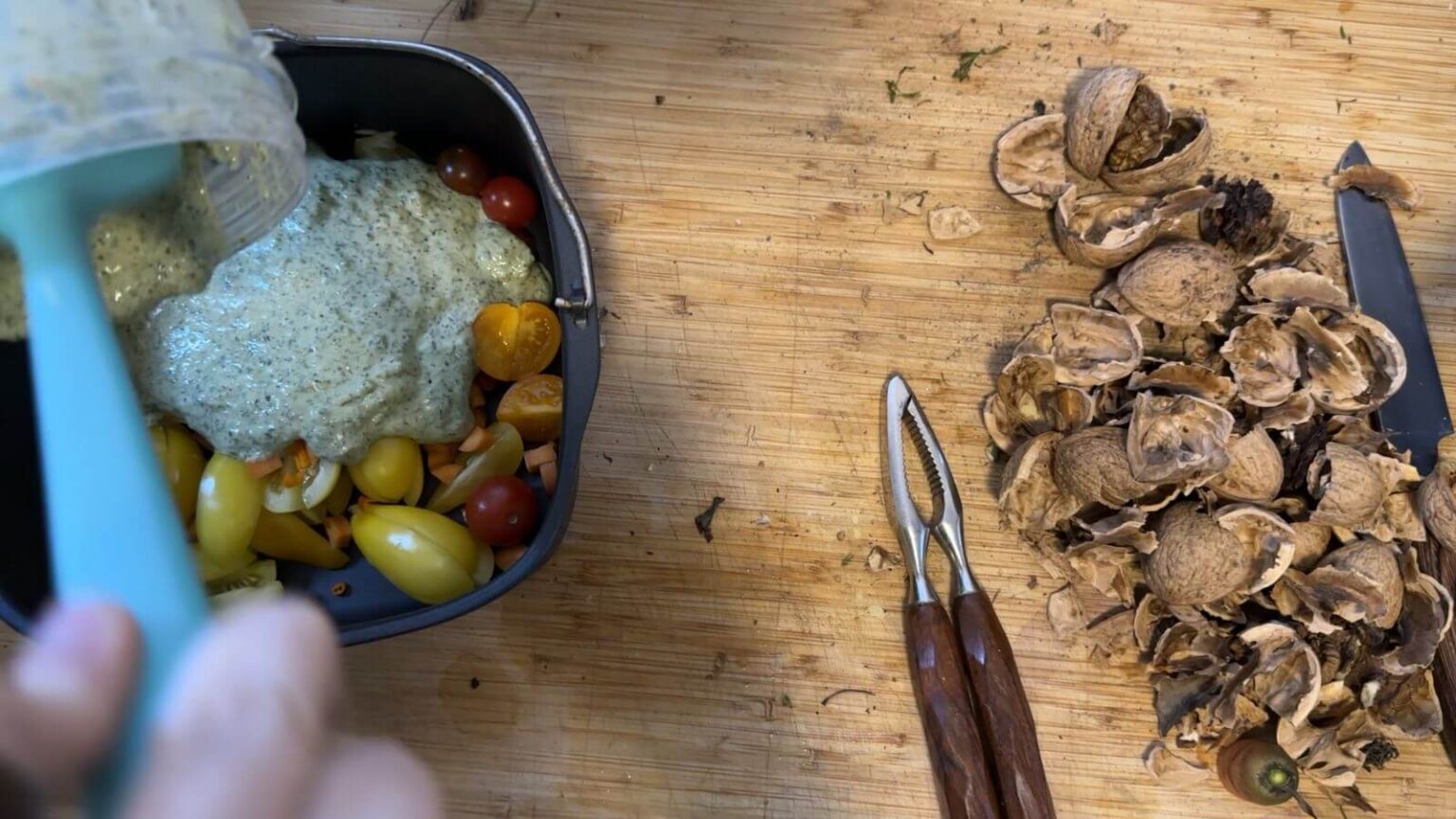 A view from above onto a wooden counter top. A pile of walnut shells is next to a nut cracker. On the left, Kate pours pesto sauce onto potatoes.