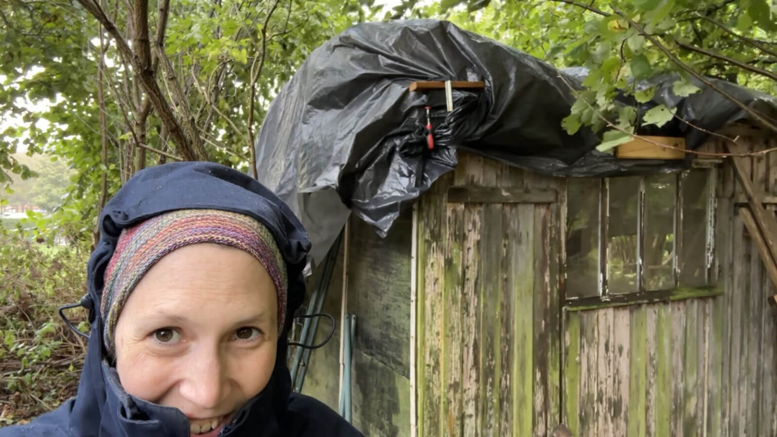 Kate is all covered up with a rain jacket hood and a head scarf. She stands in front of a shed in bad shape with a tarp over the top. The tarp is blowing in the wind.