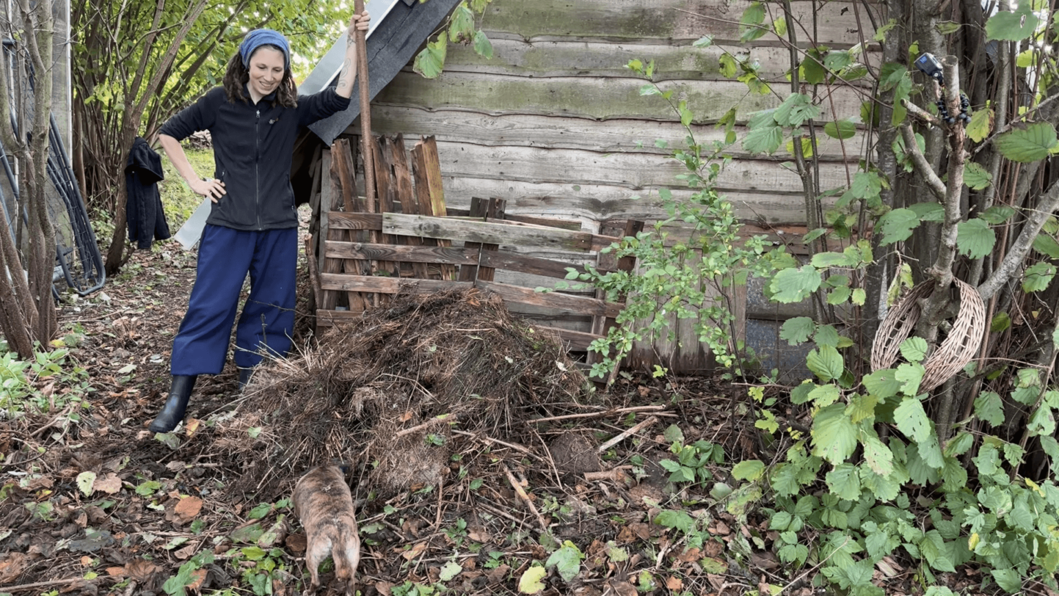 Kate stands next to a compost pile leaning on a wooden stick. Her dog is trying to dig into the pile from the front. The pile is very imperfect with larger sticks sticking out, and no side walls.