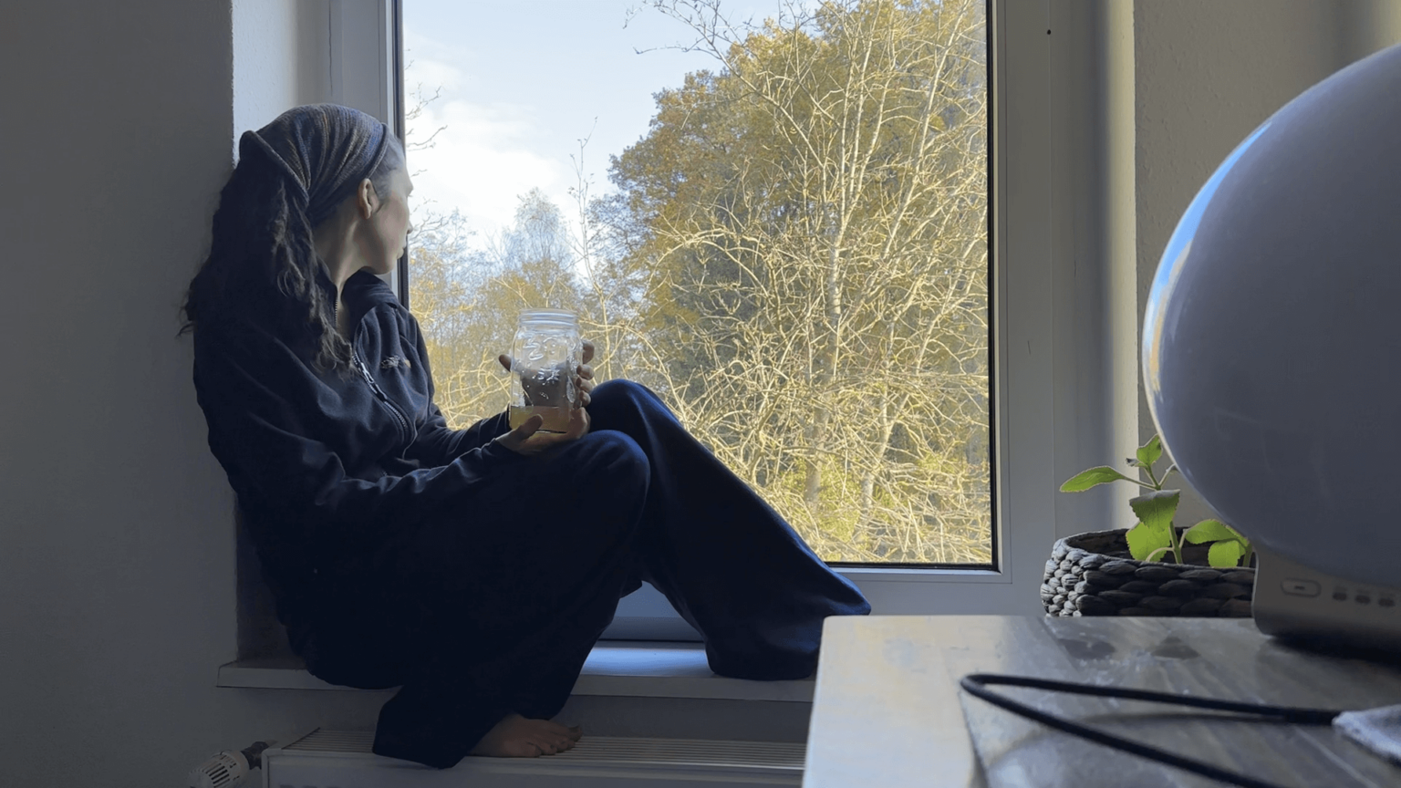 Kate sits on a windowsill, one foot on the radiator. She is holding a mason jar of kombucha in her hands, and looking out of the windows at the forest.