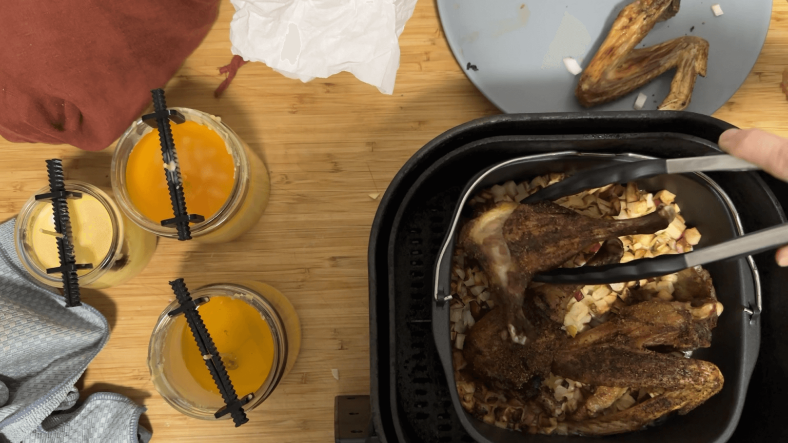 A view from above onto a bench top. On the left, three candles are drying. On the right, Kate is lifting a chicken thigh out of the air fryer.