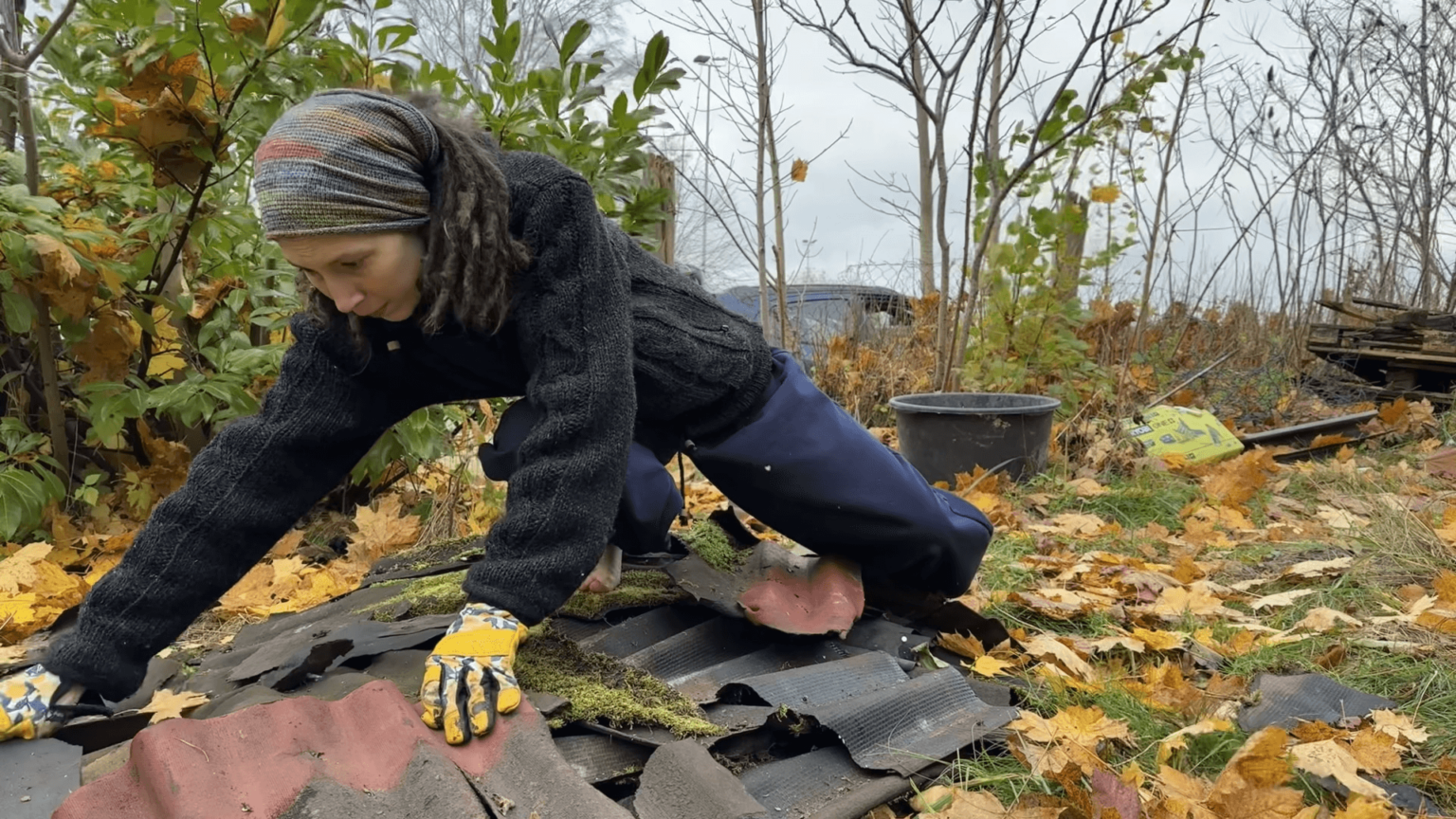 Kate leans onto a pile of old bitumen roof material. She is weaing gloves and a thick winter jacket but no shoes.