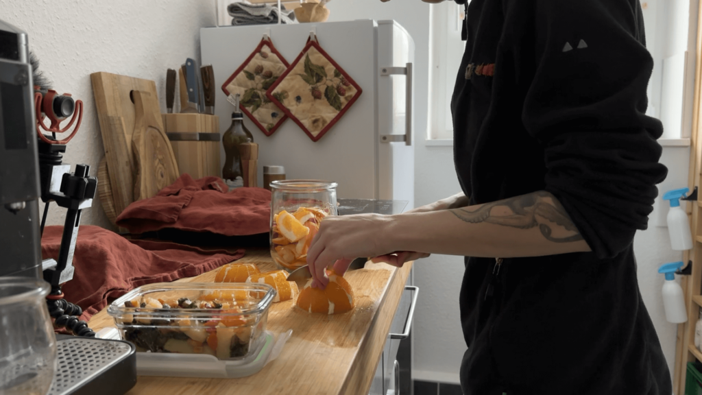 A view from the side. Only Kate's torso is visible. She is cutting an orange on her wooden bench top. The microphone stands to the side. A coffee maker is just visible in the front. Next to her are a container with chopped fruit and a jar with orange peel.