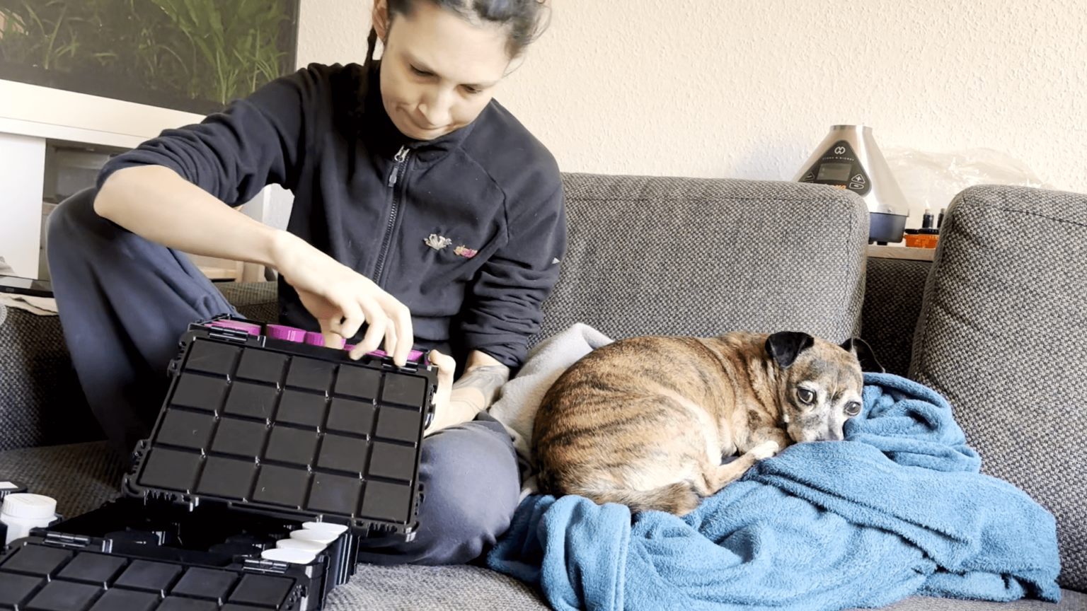 Kate sits cross-legged on a saggy gray couch. Pepper is rolled up on a turquoise blanket next to her. She is sorting through her seeds in the seed boxes she 3D-printed herself. Each box is black with a grid pattern. Within the grid sit the bottles holding the seeds. Those are in various colors.