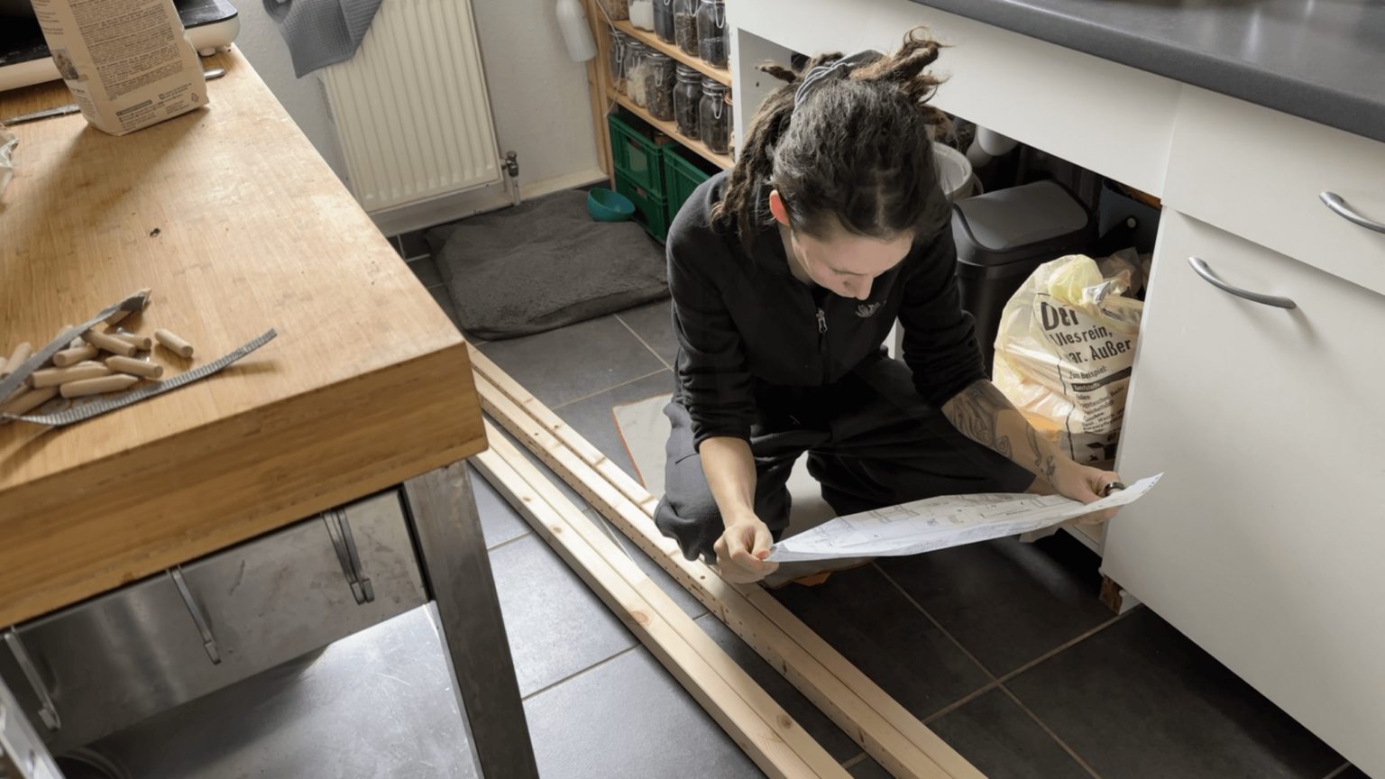 Kate squats on her kitchen floor looking at a manual for building a shelf. Next to her are some wooden plants with pre-drilled holes.