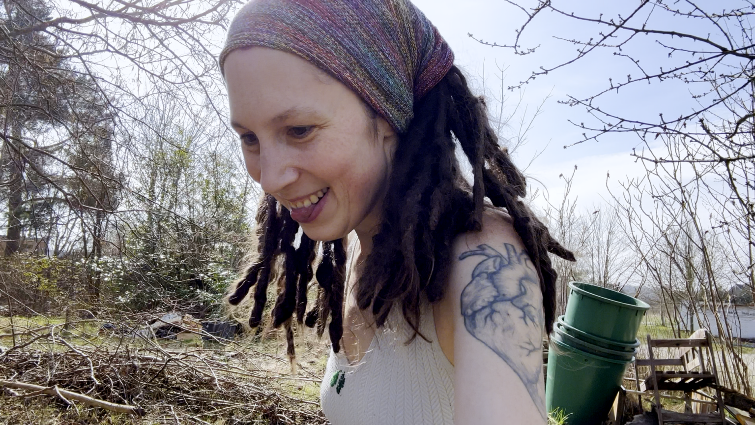 Kate's tongue is sticking out between her teeth. This is likely from exhaustion because she is pulling a heavy cart. The cart is out of sight, only the posture indicates this. Behind her is a pile of branches and some trash piles.