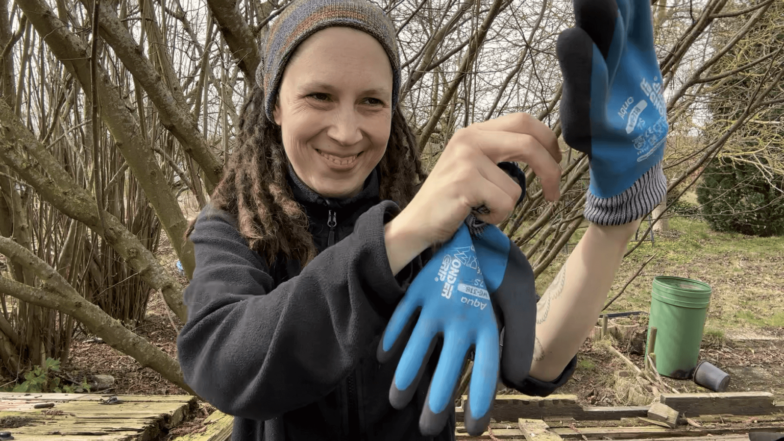 Kate grins while putting on work gloves. Behind her is a large hazelnut tree. The edge of the roof she is sticking out of is just visible toward the bottom of the screen.