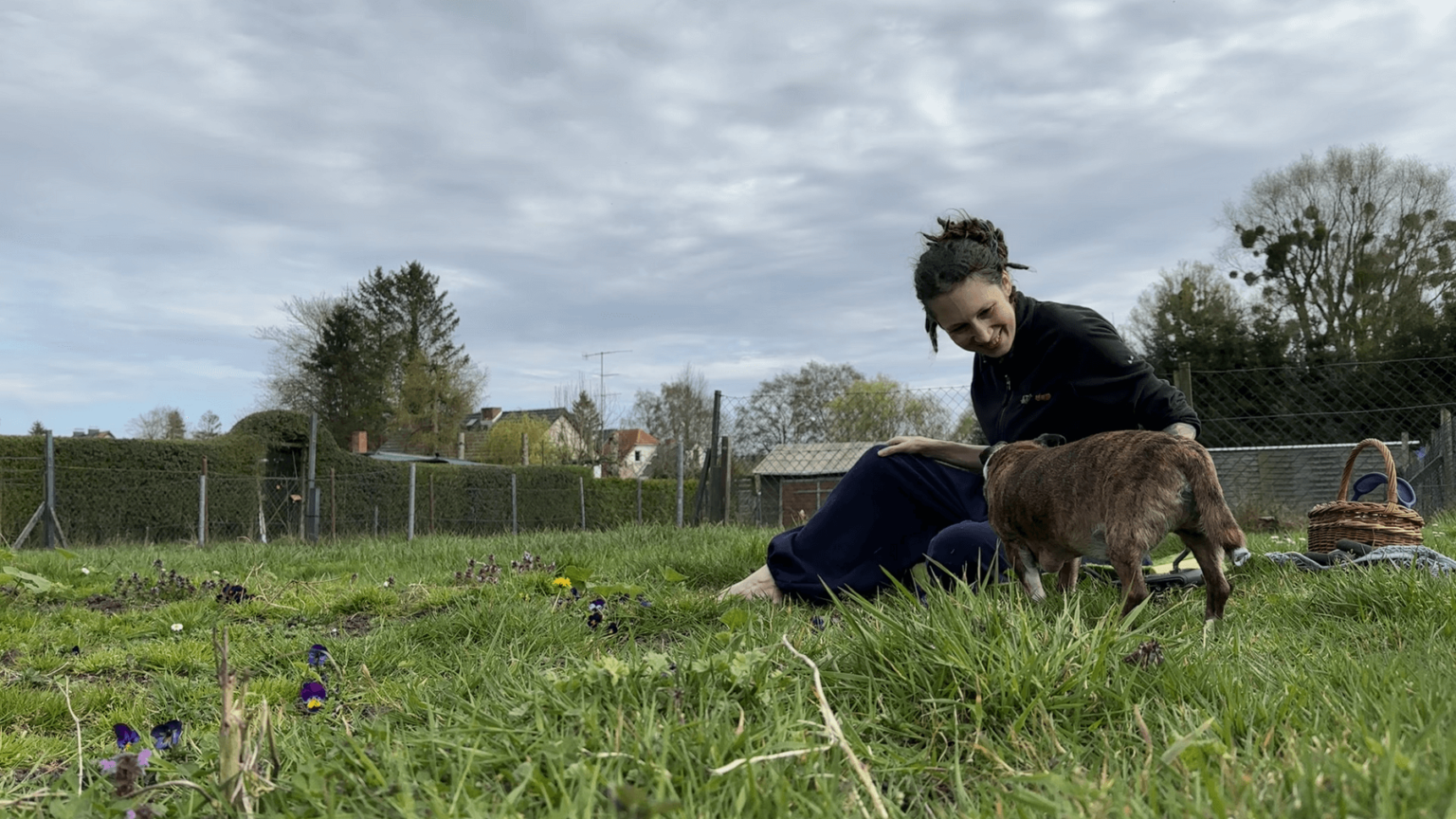 Kate sits on the grass in her garden. It is hard to see where the future bed begins and the grass ends. She is smiling while petting her dg Pepper.