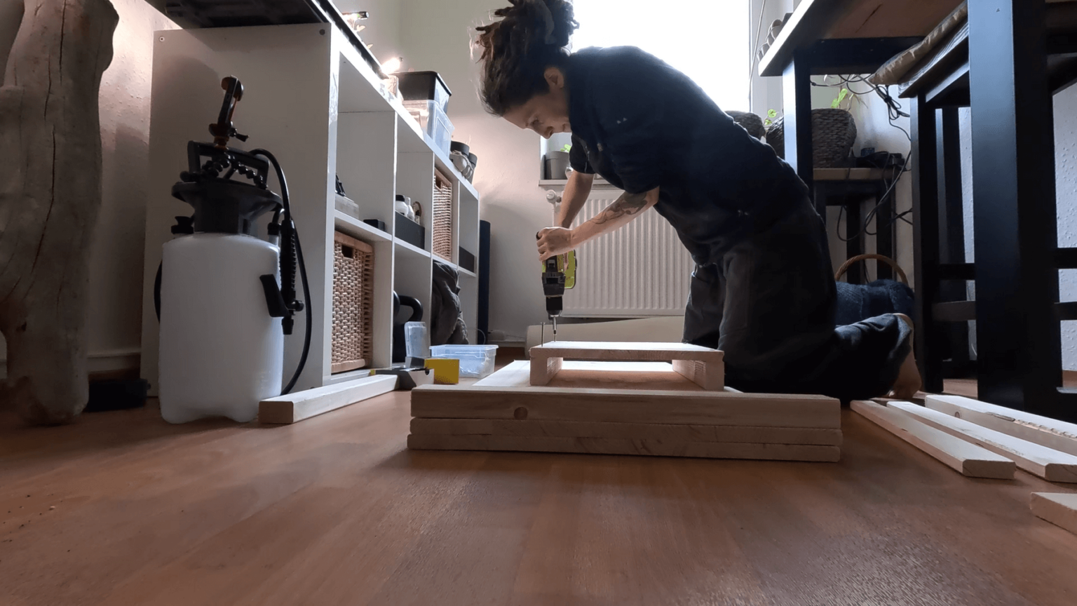 Kate kneels on top of a stack of boards with a drill. She is screwing two short beams onto a small board that will be the side of her storage box.