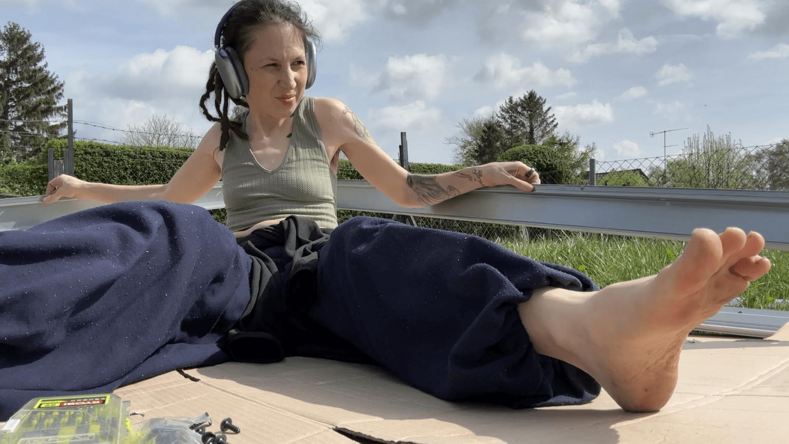 Kate is holding the bottom frame of a metal greenhouse, straining. Her legs and feet are pointing into the frame. There are headphones on her head.